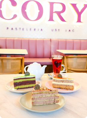 Tres rebanadas de pastel en platos, una tetera blanca y un vaso de té rojo están dispuestos sobre una mesa en una cafetería de colores pastel con asientos rosas y grandes letras en la pared de fondo.