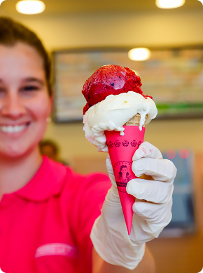 Una persona sonriente con camisa rosa y guante blanco sostiene un cucurucho de helado con una bola de helado rojo y otra de helado blanco en un envoltorio rosa. El fondo está suavemente difuminado.
