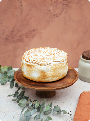 Una tarta redonda con cobertura de merengue tostado sobre un soporte de madera, con hojas verdes de eucalipto cerca y un tarro y una servilleta de fondo sobre una pared marrón.