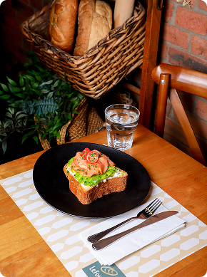 Una rebanada de pan con lechuga, salmón ahumado y tomates cherry se sirve en un plato negro con un vaso de agua, cubiertos y una cesta de pan al fondo sobre una mesa de madera.
