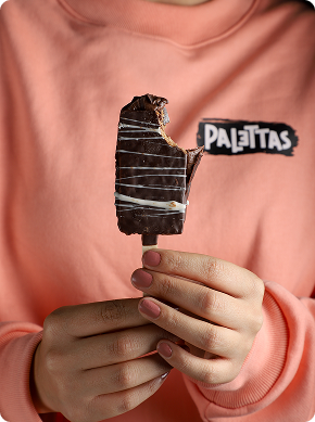 Una persona con una sudadera de color melocotón sostiene una barra de helado cubierta de chocolate mordida en un palo. La sudadera lleva el logotipo de Palettas. Las uñas de la persona están cuidadas en un tono rosa claro.